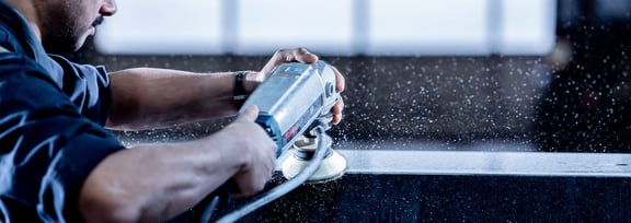 Mitarbeiter schleift mit Handschleifmaschine eine nasse Granitkante – Wassernebel sprüht während der Bearbeitung auf